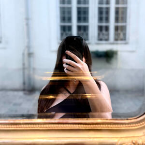 A photograph in the style of a selfie of a woman hold a phone in front of her face at a mirror. She has long dark brown hair and a black tank top on, and the wall of a building is behind her.